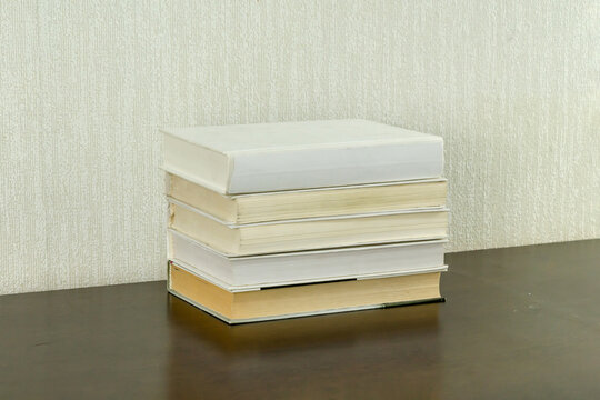 Stack of hardcover books with white and cream spines on dark wooden table against beige wall. Education, reading, literature, knowledge, study, academic, library, learning concept