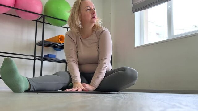 middle aged woman sits on a gym mat after exercise, taking a short rest between workouts. She adjusts her hair, exhales calmly and prepares for the next movement, showing a natural recovery moment.