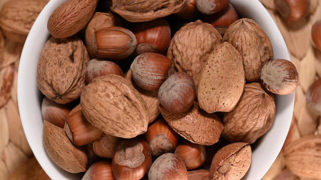 Top view of a white bowl filled with mixed nuts in their shells rotating on a wicker background