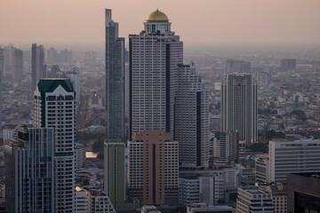 Obraz premium Panoramic aerial view of Bangkok's sprawling urban landscape at dusk, showcasing numerous modern skyscrapers and high-rise buildings, including the distinctive State Tower with its golden dome. Hazy