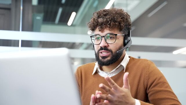 Confident businessman in a headset talking on a video call using laptop computer sitting in office. Manager has a remote business meeting. Male professional communicates at online conference. Close up