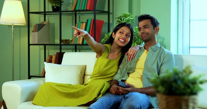Indian asian young couple watching TV with remote on lazy weekend sitting on sofa in modern Indian home while relaxing and enjoying entertainment together in a comfortable living room setup