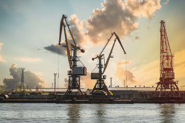 Obraz premium Industrial cranes at harbor pier during sunset. Logistics loading equipment in shipping port. Heavy machinery for freight transportation. Global trade and maritime infrastructure concept.