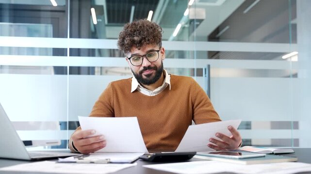 Disappointed businessman having difficulty with paperwork sitting at desk at workplace in business office. Puzzled confused accountant looks at documents in his hands and cannot understand problem