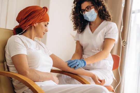 Female patient with headscarf receiving chemotherapy treatment from nurse in a hospital room, IV drip stand and natural light visible through window