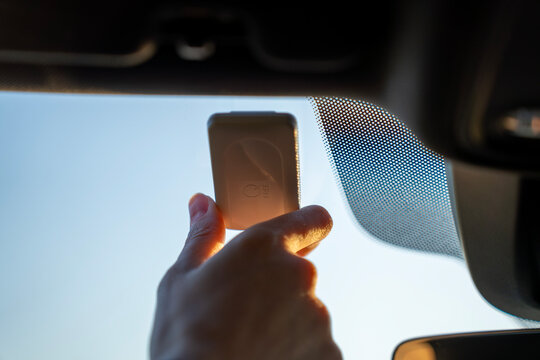 Hand holding toll transponder mounted on sun visor in modern car interior, close-up of electronic toll payment device near windshield.