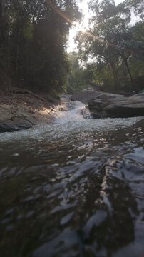 Slow motion of a peaceful stream flows over rocks in a forest, with sunlight filtering through the trees, creating a serene and tranquil atmosphere.