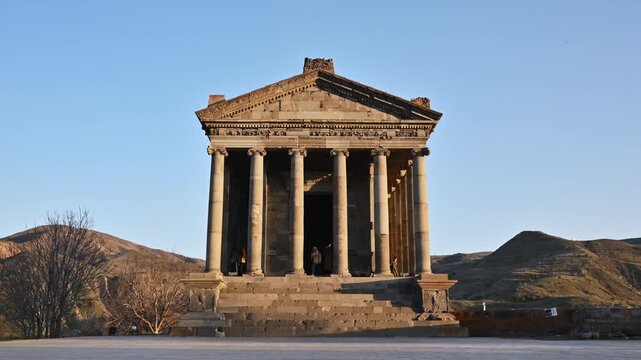 Ancient temple stands tall under clear blue sky. Stone columns support grand roof structure. Hills surround the site. One person walks near entrance steps