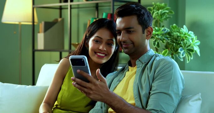 Indian asian young couple surfing smartphone together sitting on sofa in modern Indian home on lazy weekend while looking at screen and sharing digital content in a bright living room setup