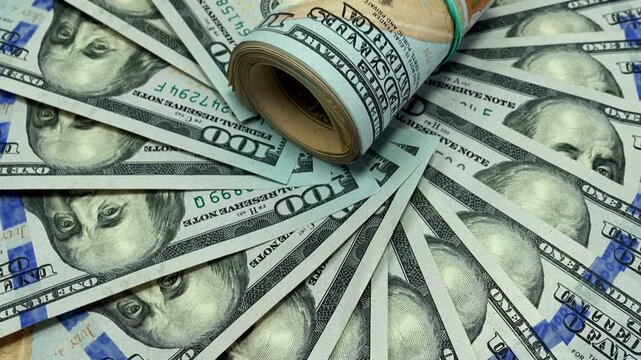 Close-up of fanned US hundred dollar bills while a rolled bundle of cash moves into view on top of the money spread