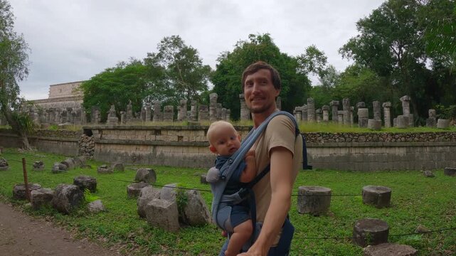 Father walking with little son in ergo carrier along Temple of the Thousand Columns colonnade in Chichen Itza, Mexico. Selfie slow motion travel video. Family bonding and tourism concept.
