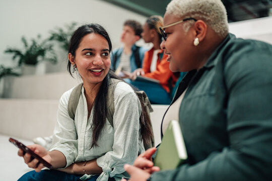 Two diverse female students interacting and discussing on a university staircase while holding a smartphone and a notebook