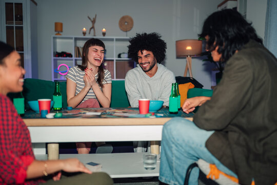 Diverse group of young adult friends playing a board game, drinking beverages, and having fun together in a living room during an evening gathering