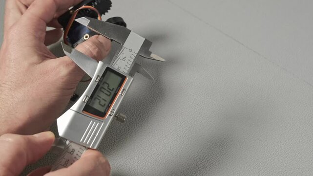 An engineer works with a digital caliper to measure a small robotic servo component with gears and wiring. Taking size, wide, length The precise measurement is shown on the LCD screen of the tool.