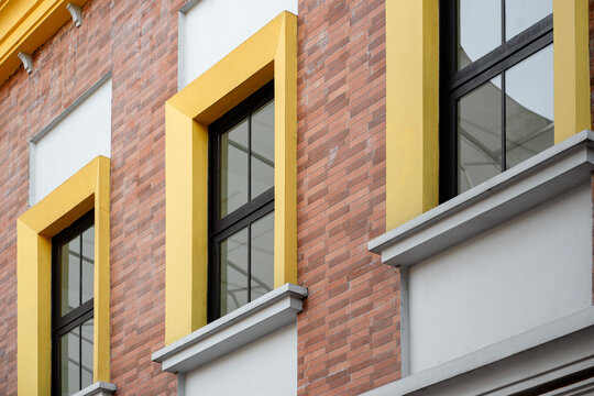 Close-up architectural detail of a modern building exterior featuring a textured red brick wall and black-framed windows enclosed by vibrant, thick yellow borders. The contrast between the classic bri