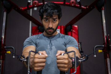 Obraz premium Young adult man doing intense workout at gym using cable machine displaying deep determination and focused emotional expression while building physical strength during fitness exercise