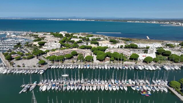 Wide aerial view of Grau du Roi France with dense Port Camargue marina full of sailboats in long rows white buildings lush trees and vast blue sea under clear sky