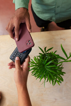 Two people exchanging maroon phone and black numeric-keypad phone on wood table comparing cameras