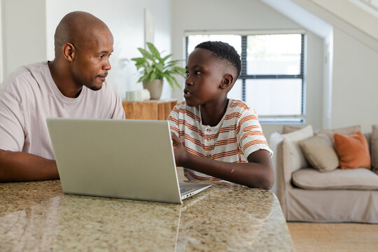 African American father and adolescent son sitting at granite counter, using open silver laptop