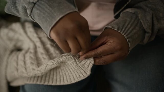 Cropped close up handheld shot of hands of unrecognizable Black woman sewing button on grey knitted cardigan, fixing clothes to save money during financial hardship