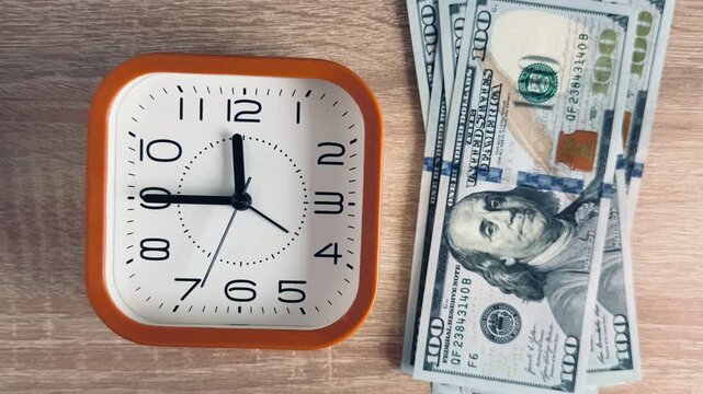 Clock hands spinning fast next to growing stack of US hundred dollar bills on wooden table, time is money concept, stop motion