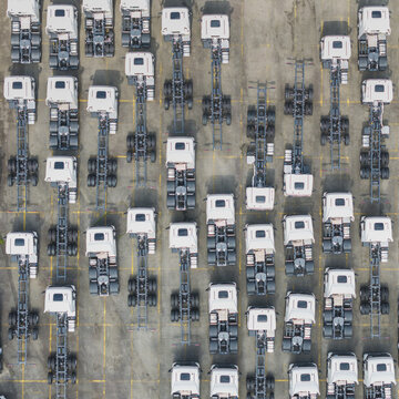 Aerial view of rows of white semi-truck chassis parked in a logistics lot with yellow grid markings on the asphalt, Taiwan.