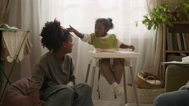 Handheld pan shot of caring Black mother repairing clothes on couch while watching teenage daughter playing with little sister in high chair, sharing small low income family apartment together