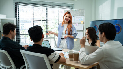 Fototapeta premium young Asian Gen Z business team meeting and collaborate in modern office. Creative woman with orange hair discussing project strategy with colleagues. Concept of startup and youth empowerment.
