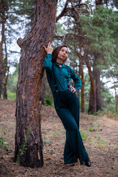 Woman in teal outfit leaning on tree in pine forest