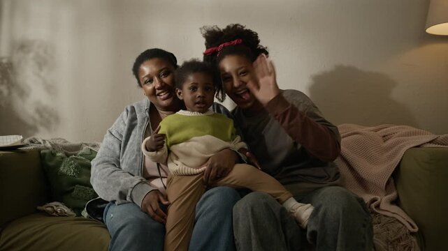 Medium long handheld portrait of loving Black family consisting of single mother with toddler and teenage daughters waving at camera on couch in dimly lit modest apartment
