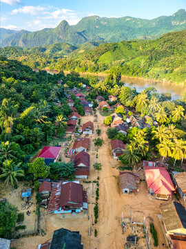 Aerial view of a small village with red-roofed houses along a dirt road surrounded by lush tropical greenery and karst mountains by the Nam Ou River in Nong Khiaw, Luang Prabang Province, Laos.