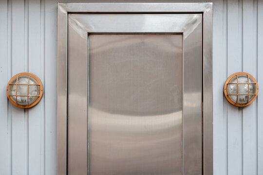 Stainless steel door on a grooved white wall flanked by two industrial round bulkhead wall lights. Modern architectural exterior detail.
