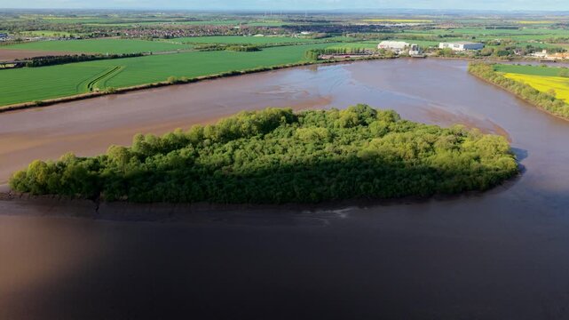 Aerial drone view of Howden Dyke Island surrounded by the brown muddy estuary water of the River Ouse and a green rural landscape in sunny conditions near Goole East Yorkshire UK