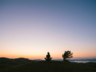 Fototapeta premium Serenity in Silhouette: A lone figure finds inner peace, meditating atop a serene hill under a vast, colorful sky, embracing tranquility and self-reflection.