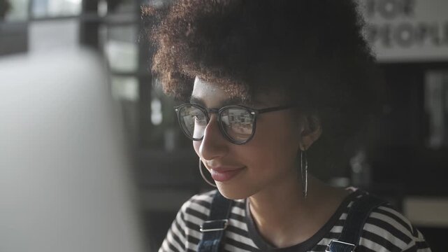Close-up portrait of a focused young woman with afro hairstyle and glasses looking at computer screen in a modern interior. Stylish casual outfit, soft natural light and calm concentrated expression