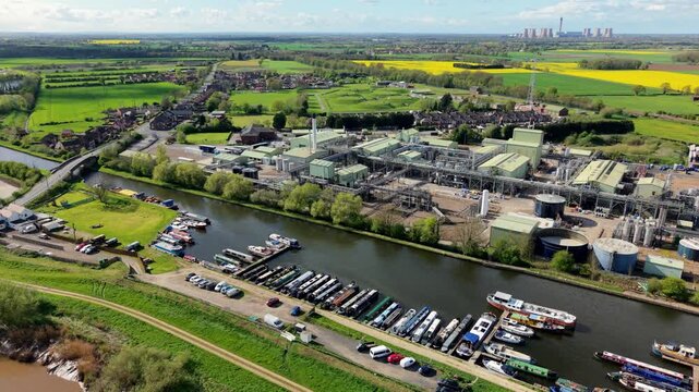 Aerial view of industrial chemical production factory on river bank with narrowboats docked and power station towers in the background and sunny rapeseed fields UK