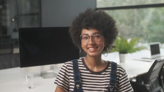 Portrait of a smiling young woman with afro hairstyle and eyeglasses looking at camera in a modern office interior. Friendly female professional in striped shirt and denim overalls posing at workplace