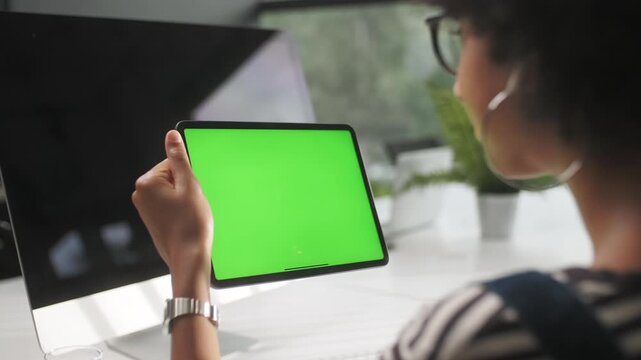 Rear side view of a young woman holding a tablet with chroma key green screen and swiping upward in a modern office. Female user interacts with touchscreen device while sitting at desk near computer