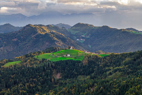 Aerial view of St. Thomas's Church on a vibrant green hilltop surrounded by dense autumn forests and misty mountain peaks in Ratece Planica, Jesenice, Slovenia.