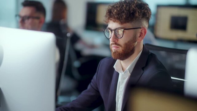Concentrated young businessman in glasses working at computer in modern open plan office. Professional male employee in formal suit focused on screen while coworkers work in blurred background