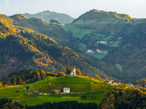 Aerial view of St. Thomas's Church on a lush green hilltop surrounded by dense forests and rolling alpine mountains in Ratece Planica, Jesenice, Slovenia.