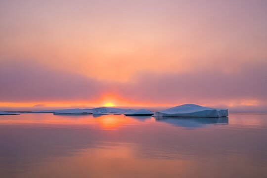 Breathtaking arctic sunset paints the horizon with vibrant orange and purple hues, beautifully reflecting on calm water as large icebergs float.