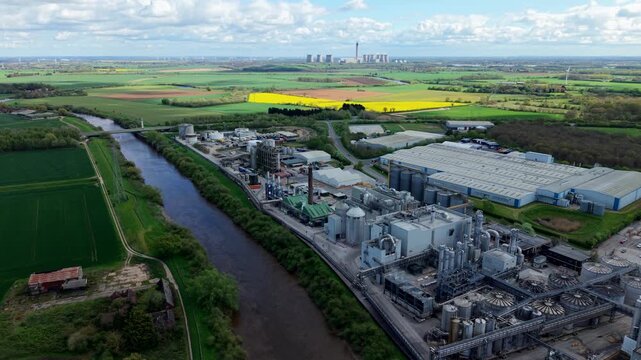 Drone shot of a large production plant complex featuring vertical storage silos and pipelines next to a river