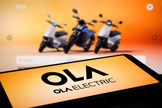 Ola Electric logo on smartphone screen with electric scooters lineup in background, Indian EV revolution and electric vehicle startup brand concept for stock market investors