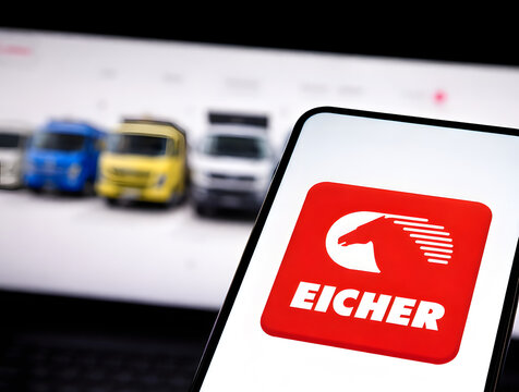 Eicher Motors brand logo displayed on smartphone screen with commercial trucks lineup on company website in background, Indian automobile and commercial vehicle sector concept