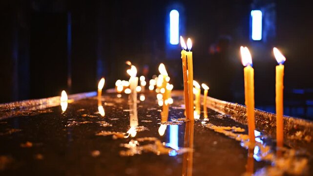 Candles burn softly in a dim, sacred space. Warm light reflects on wet, dark surface. Background shows faint blue window light. Candles vary in height and flame intensity. Scene evokes quiet reverence