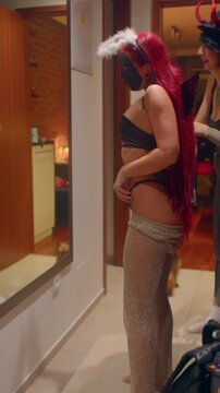 friends in hallway adjusting costumes in front of mirror, halo and devil horn accessories, red hair and fitted outfits, stylist assisting with fitting, playful anticipation before night out Vertical