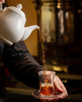 Close-up of pouring hot black tea from a white ceramic teapot into a traditional armudu glass. Authentic tea culture with a samovar in the background.