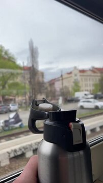Vertical stock video hand holding bottle near train window, stainless thermos with flip lid, commuter inspecting lock, finger pressing button for sip, urban cityscape passing