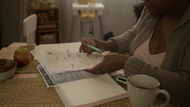 Handheld shot of young African American mother reviewing utility bills and marking overdue payments at table, then placing her toddler daughter on laps, showing financial struggle in loving family